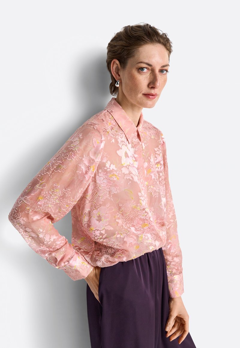 Woman wearing a sheer pink floral blouse and dark high-waisted pants, standing with one hand in pocket against a plain light background.