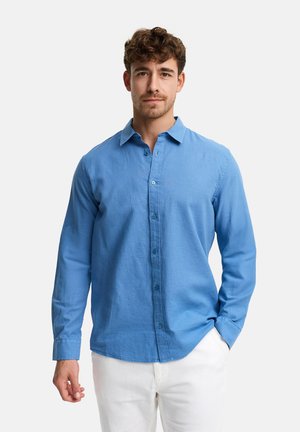 Młody mężczyzna o kręconych włosach, ubrany w jasnoniebieską koszulę na guziki i białe spodnie, stojący na tle jednolitego białego tła.