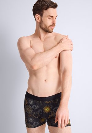 Homme torse nu portant un boxer noir avec des motifs de soleil, tenant son épaule gauche et regardant vers le bas sur un fond clair uni.
