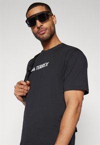 Sort bomulds T-shirt med "TERREX" logo i hvid, parret med store sorte solbriller og en sort skuldertaske.