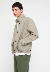 Jasnobeżowa kurtka z kołnierzem typu stójka, czarnymi guzikami, dwoma przednimi kieszeniami i luźnym krojem, zestawiona z oliwkowozielonymi spodniami.