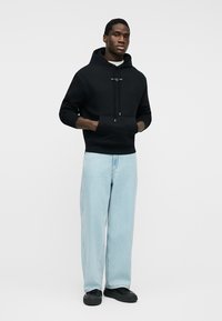 Férfi áll, kezét egy fekete kapucnis pulóver zsebében tartja, világoskék, széles szárú farmert és fekete cipőt visel, egyszerű háttér előtt.