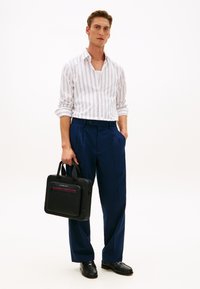Άνδρας που φοράει άσπρο πουκάμισο με ρίγες, μπλε παντελόνι, μαύρα μοκασίνια, κρατώντας μαύρη τσάντα χαρτοφύλακα σε λευκό φόντο.