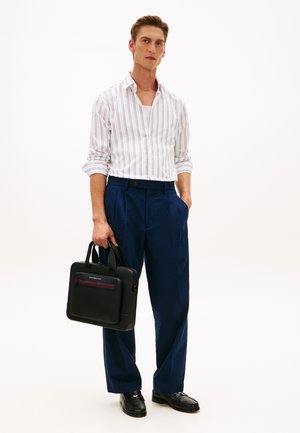 Homme portant une chemise à rayures blanches, un pantalon bleu, des mocassins noirs, tenant une mallette noire sur fond blanc.