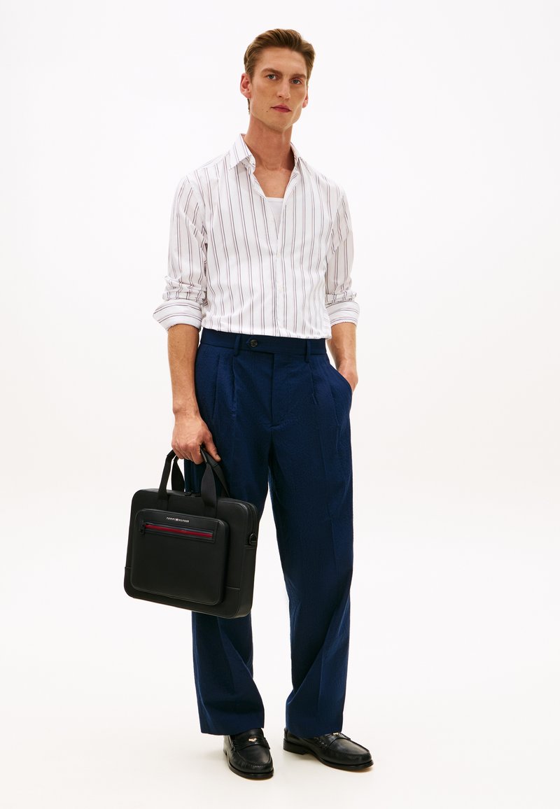 Άνδρας που φοράει άσπρο πουκάμισο με ρίγες, μπλε παντελόνι, μαύρα μοκασίνια, κρατώντας μαύρη τσάντα χαρτοφύλακα σ ε λευκό φόντο.