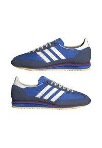 Paire de baskets Adidas bleues avec des bandes blanches, des accents en daim gris, des lacets blancs et des semelles jaunes, présentées côte à côte sur fond blanc.