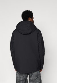 Άνδρας γυρισμένος προς τα πίσω, φορώντας μαύρο μπουφάν με κουκούλα και γκρι καρό παντελόνι, σε έναν απλό ανοιχτό φόντο.