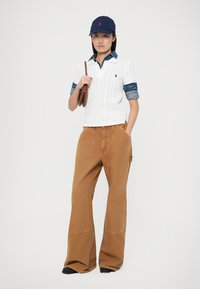 Weiße Kabelstrick-Polohemd über einem Denim-Hemd, kombiniert mit weit geschnittenen braunen Hosen. Eine marineblaue Mütze mit rotem Logo und eine braune Tasche runden den Look ab.