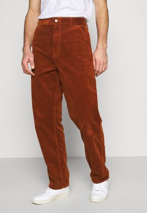 Mand iført rustfarvede kordfløjlsbukser, hvide sneakers og en hvid t-shirt, stående mod en ensfarvet baggrund.