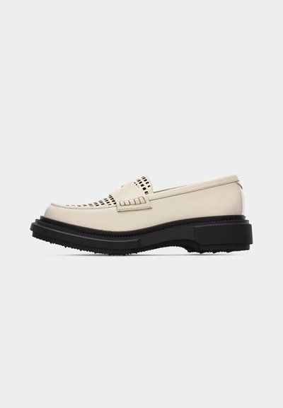Mocassin en cuir beige avec semelle noire épaisse, empeigne perforée et détail de sangle décorative, présenté de profil sur un fond blanc.