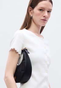 Δομημένη μαύρη δερμάτινη τσάντα με κυρτό σχήμα και χρυσά εξαρτήματα, συνδυασμένη με λευκό, υφασμάτινο τοπ με κυματιστά μανίκια.