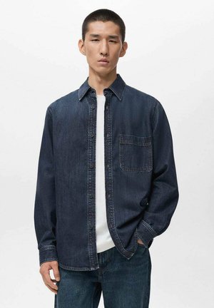 Donkerdenim overhemd met knopen en een puntige kraag, een enkele borstzak en een relaxte pasvorm. Gekozen over een effen wit T-shirt.
