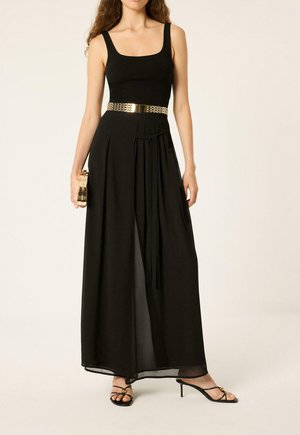 Femme portant une robe maxi noire sans manches avec une ceinture dorée, tenant une pochette dorée et portant des sandales à talons noires.