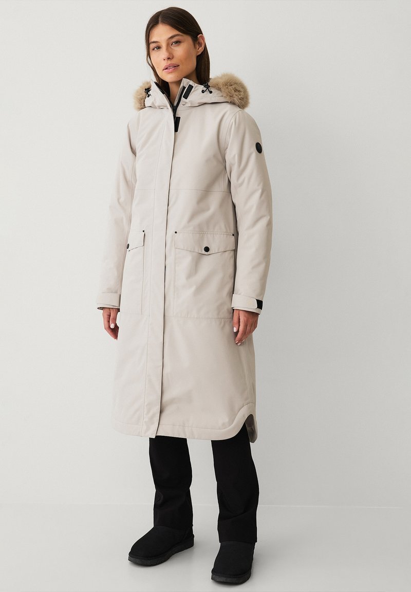 Lungo cappotto imbottito beige con cappuccio bordato di pelliccia, chiusura con zip, due grandi tasche e dettagli neri sui polsini e sul colletto. Indossato con pantaloni e stivali neri.