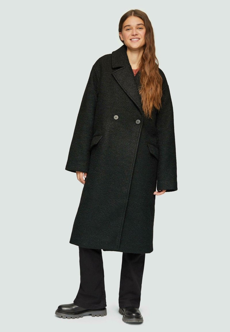 Zwarte wolblend jas met een dubbelgebreide ontwerp, grote revers, zijzakken en een oversized pasvorm. Gecombineerd met donkere broek en chunky laarzen.