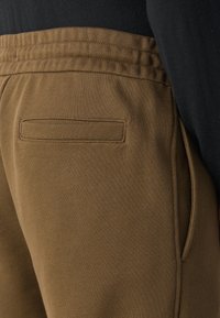 Braune Jogginghosen aus einem strukturierten Stoff. Mit elastischem Bund, seitlicher Tasche und einer hinteren aufgenähten Tasche mit schlichten Nähten.