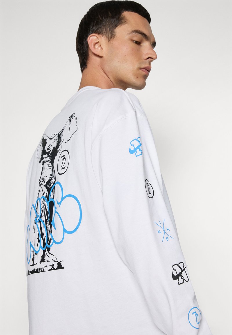 Chemise à manches longues blanche avec un design graphique d'une statue et des accents colorés. Comprend des logos bleus et noirs sur les manches.