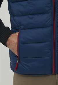 Mornarsko puffer telovnik z vodoravnimi prešitimi odseki, rdečimi zadrgami in stranskimi žepi. Nošen preko črnega pletenega puloverja.