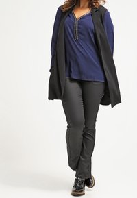 Gilet senza maniche nero sopra una blusa blu navy con scollo decorativo, abbinato a pantaloni neri attillati e sneakers nere con suola texturizzata.