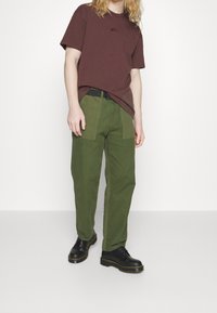 Hnědé bavlněné tričko s malým logem, kombinované se zelenými širokými kalhotami s předními kapsami a černými šněrovacími botami se žlutým stehem.