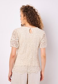 Blusa de encaje en color blanco roto con mangas cortas y abullonadas. Presenta un dobladillo con forma de ondas y una abertura en forma de llave en la parte posterior, que muestra patrones intrincados.