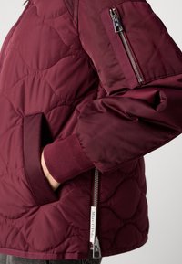 Giacca trapuntata bordeaux con colletto alto, tasche laterali e tasca sulla manica con zip; presenta polsini elasticizzati e texture del tessuto liscia.