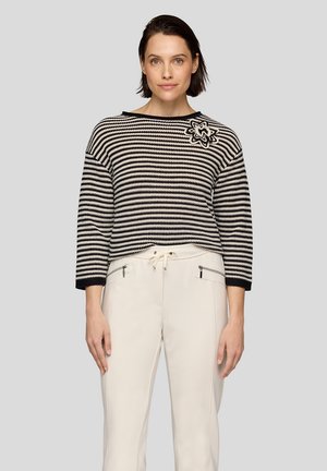 Gestreifter Pullover in Schwarz-Weiß mit Dreiviertelärmeln und einem Blumenpatch auf der linken Schulter, kombiniert mit hellen Hosen.