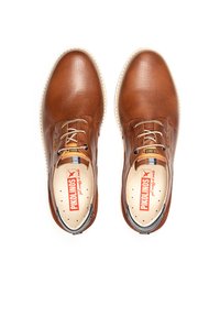 Zapatos de cuero marrón con textura perforada, punta redonda y diseño de cordones. Cuentan con una suela blanca y una etiqueta interior roja.