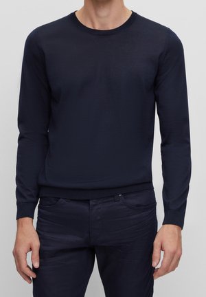 Mand iført en tætsiddende mørkeblå langærmet sweater og matchende mørkeblå bukser, stående mod en ensfarvet grå baggrund.