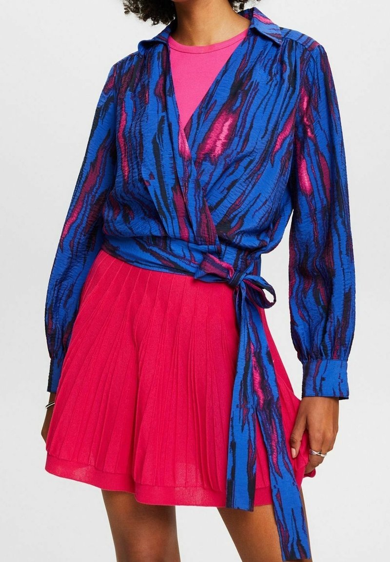 Blouse cache-cœur à motifs bleus et roses avec manches longues, nouée à la taille, associée à une jupe plissée rose, créant une tenue éclatante.