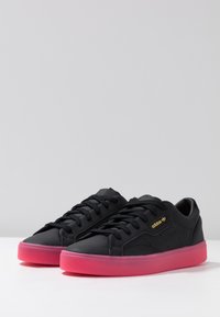 Black leather sneakers with a smooth texture, flat black laces, gold logo, and a vivid pink translucent sole. Classic sports design.