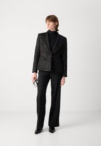 Zwarte blazer met bloemenpatroon en een gladde textuur, gecombineerd met zwarte broek en glanzende zwarte schoenen. Houdt een zilveren metalen clutch vast.