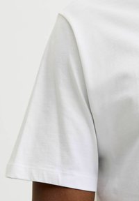 Prim-plan al brațului unei persoane îmbrăcate cu un tricou din bumbac alb, cu mânecă scurtă, pe un fundal neutru.