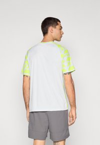 Camiseta atlética blanca de manga corta con patrones geométricos en amarillo neón en las mangas. Hecha de una tela ligera y transpirable.