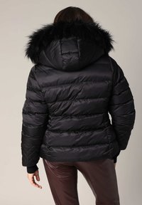 Doudoune noire avec capuche présentant une bordure en fausse fourrure, texture matelassée et poignets côtelés. Le dos met en avant un design ajusté.