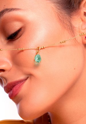 Goldkette mit runden Perlen und einem grünen Tropfen-Edelstein-Anhänger, auf der Haut positioniert, um das filigrane Design des Schmuckstücks zu betonen.