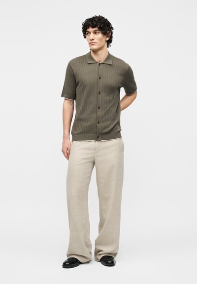 Νέος άνδρας στέκεται με τα χέρια πίσω από την πλάτη, φοράει πουκάμισο με κοντά μανίκια σε ελαιί πράσινο χρώμα με κουμπιά, μπεζ παντελόνι με φαρδιά μπατζάκια και μαύρα παπούτσια.
