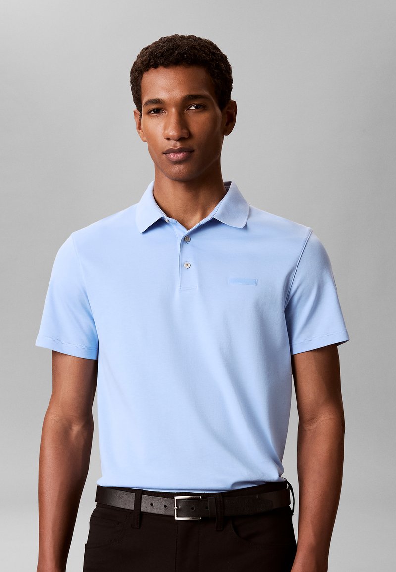 Man draagt een lichtblauw kortarmig poloshirt en een zwarte riem met zwarte broek, staand tegen een effen grijze achtergrond.