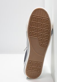 Podeszwa sneakersa wykonana z brązowej gumy z teksturowanym, zygzakowatym i geometrycznym wzorem. Boki są białe, z granatowym materiałem płóciennym powyżej.