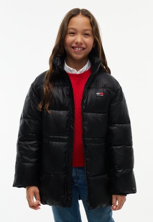 Ung person smiler, iført en sort dunjakke, rød sweater, hvid skjorte og blå jeans, står mod en ensfarvet hvid baggrund.