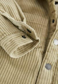 Khaki žebrovaná bunda s černými patentkami, měkkou texturou a límečkovým designem s akcentovým knoflíkovým detailem na přední straně.