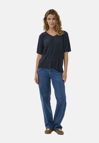 Kvinde med krøllet hår iført en sort kortærmet plisseret top, blå jeans og brune sneakers, stående mod en ensfarvet hvid baggrund.