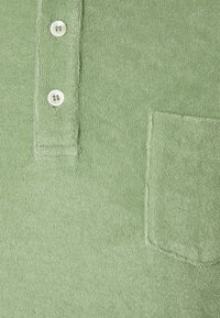 Ljusgrön frottétyg med en liten bröstficka och två vita knappar på en knappslå.