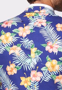 Kwiatowa marynarka w kwiaty na niebieskim tle z dużymi kwiatami hibiskusa w odcieniach różu, żółci i pomarańczy, ozdobiona zielonymi liśćmi i tropikalnymi frondami.