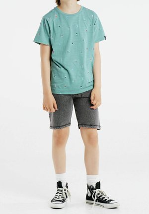 Bambino che indossa una maglietta verde con piccolo disegno stampato, pantaloni corti in denim grigi, calzini bianchi e scarpe nere alte, in piedi su uno sfondo bianco.