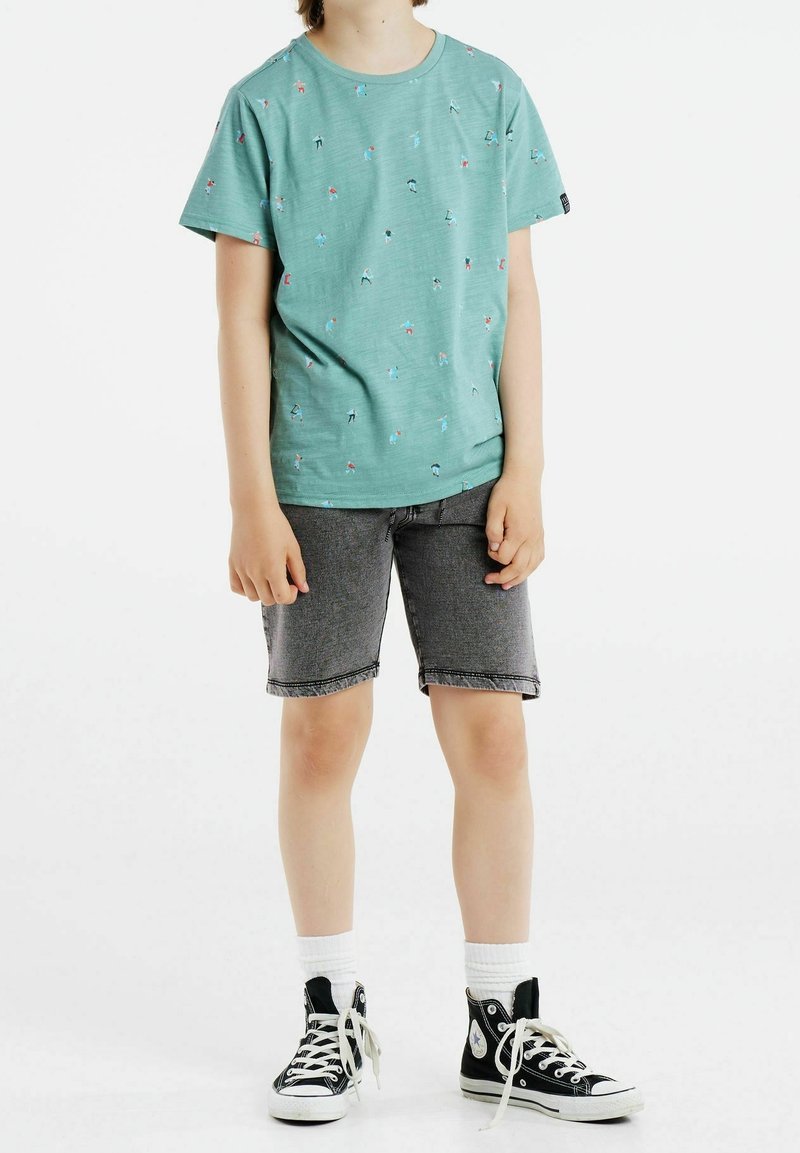 Lapsi, joka on pukeutunut vihreään t-paitaan, jossa on pieni kuvio, harmaisiin farkkushortseihin, valkoisiin sukkiin ja mustiin korkeatennareihin, seisoo valkoista taustaa vasten.