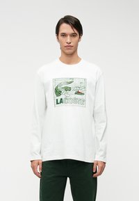 T-shirt bianca a maniche lunghe in cotone, con una grafica verde di un coccodrillo e il testo "LACOSTE" all'interno di un riquadro. Design semplice e vestibilità rilassata.