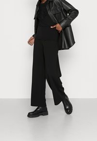 Camisa de cuero negra oversized superpuesta sobre una camiseta gráfica negra y pantalones anchos de pierna ancha negros. Botines negros de suela gruesa.