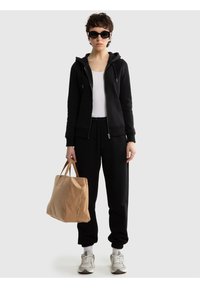 Persoană îmbrăcată cu hanorac negru, maiou alb, pantaloni de trening negri, adidași, ochelari de soare și ținând o geantă mare bej, pe un fundal simplu.