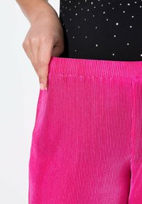 Pantalones de pana de color rosa brillante con una textura acanalada, que presentan una cinturilla elástica y un bolsillo lateral. Mano sosteniendo la cinturilla.